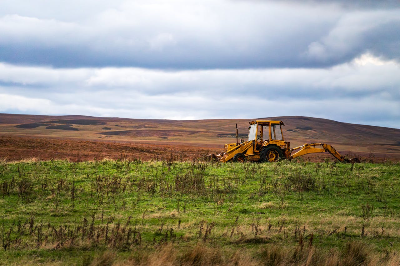 gallery-01 A solitary yellow backhoe on a wide green field against a backdrop of hills and a cloudy sky.